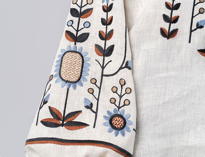 A close-up shot of a cream-colored linen blouse featuring ornate floral embroidery in blue, rust, and black tones on a voluminous puff sleeve