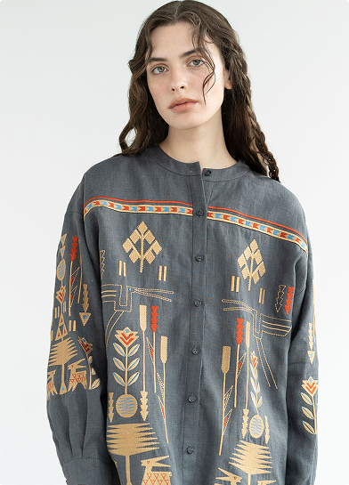 A woman with wavy brown hair wearing a grey button-down shirt featuring intricate, folk-style embroidery in gold, orange, and blue