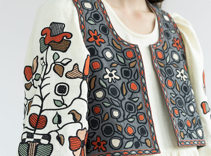 A close-up shot of a cream-colored, long-sleeved top decorated with a vertical floral vine pattern and layered with a grey open vest